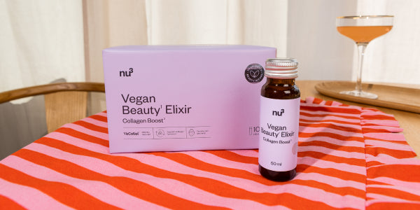 Confezione e flaconcino di Vegan Beauty Elixir su un tavolo con tovaglia a righe e bicchiere sullo sfondo